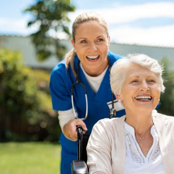 Happy nurse and elderly woman sitting in wheelchair enjoying outdoor treatment session.