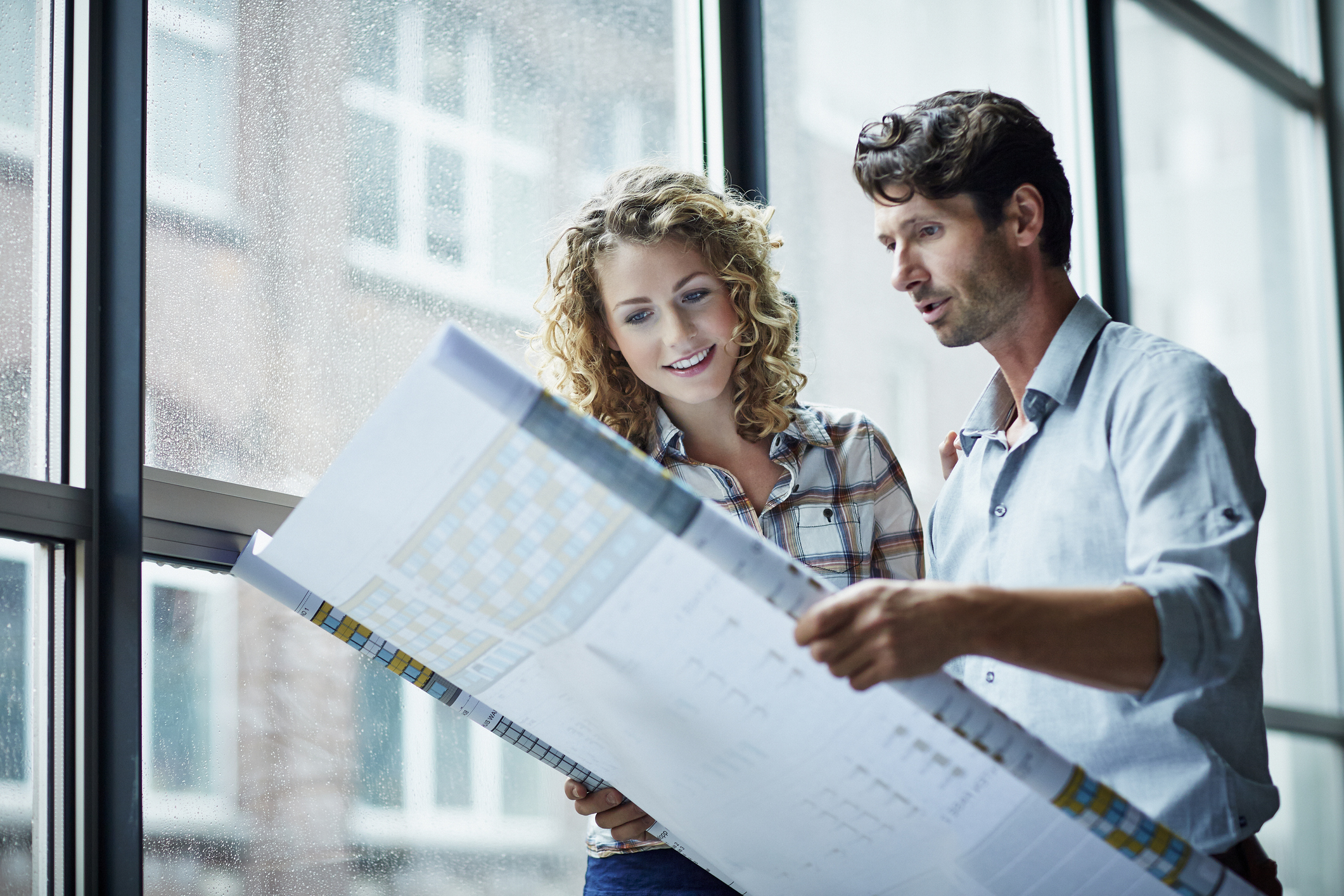 Two architects checking blueprint by window in modern studio