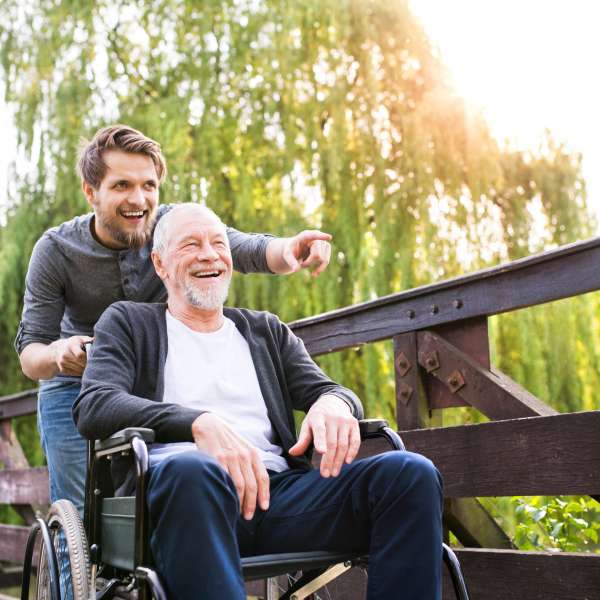 A personal aide walking with a senior man in wheelchair at park.