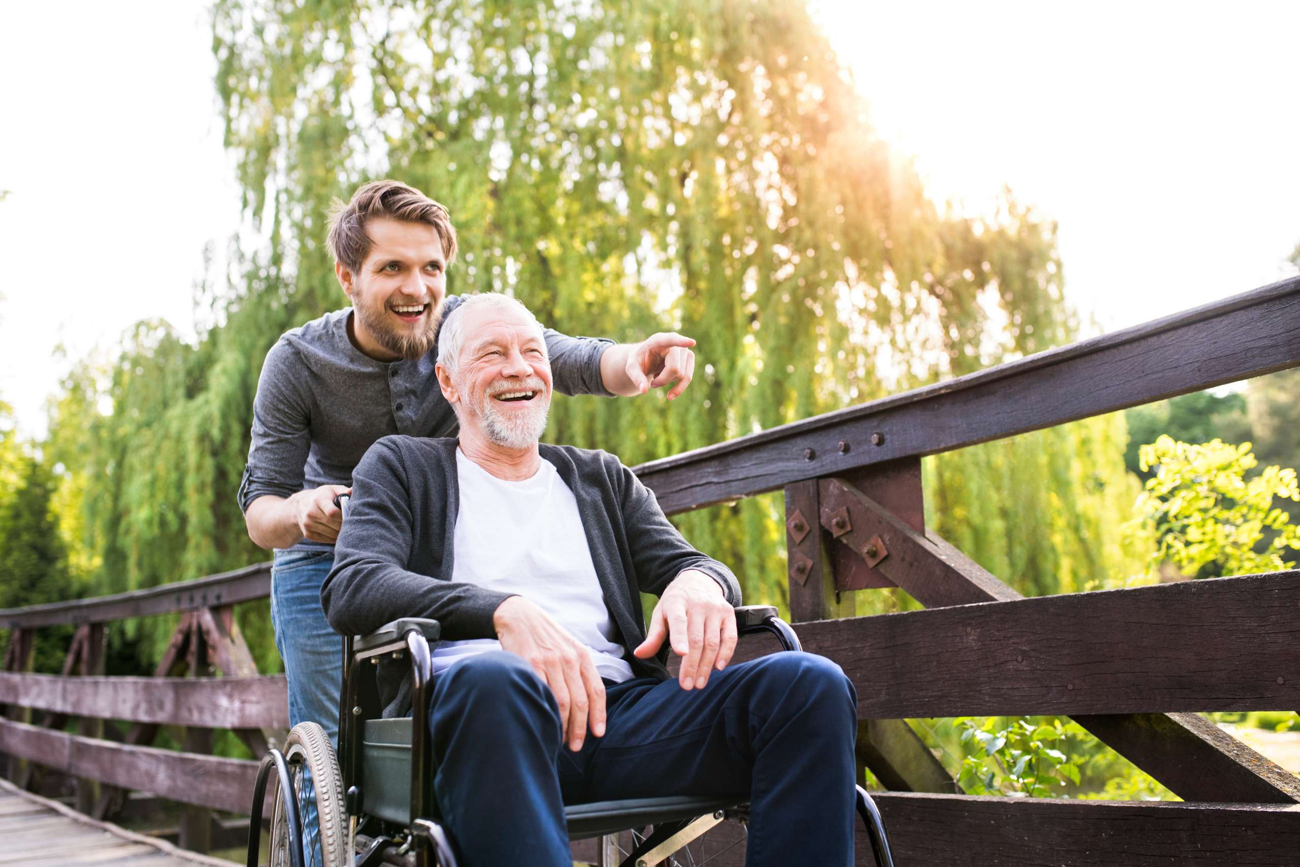 A personal aide walking with a senior man in wheelchair at park.