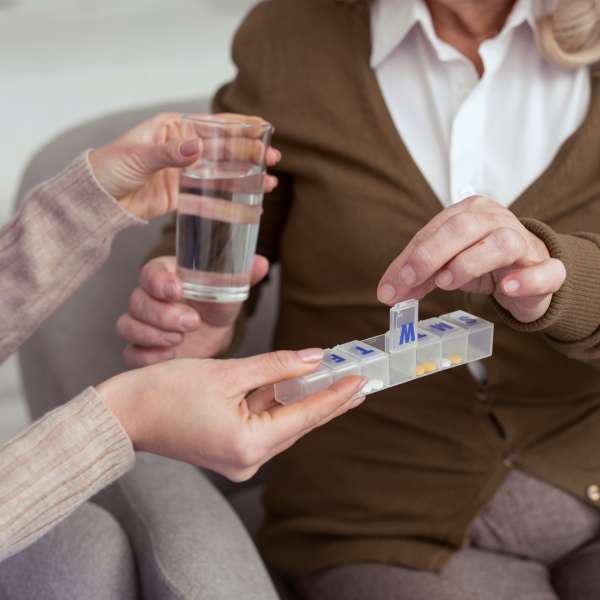 Female holding container for medication while giving glass with water to elder woman