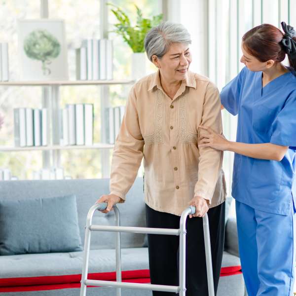 Young professional successful friendly female nurse in blue hospital uniform helping supporting physical therapy senior woman patient walking via four legs walker