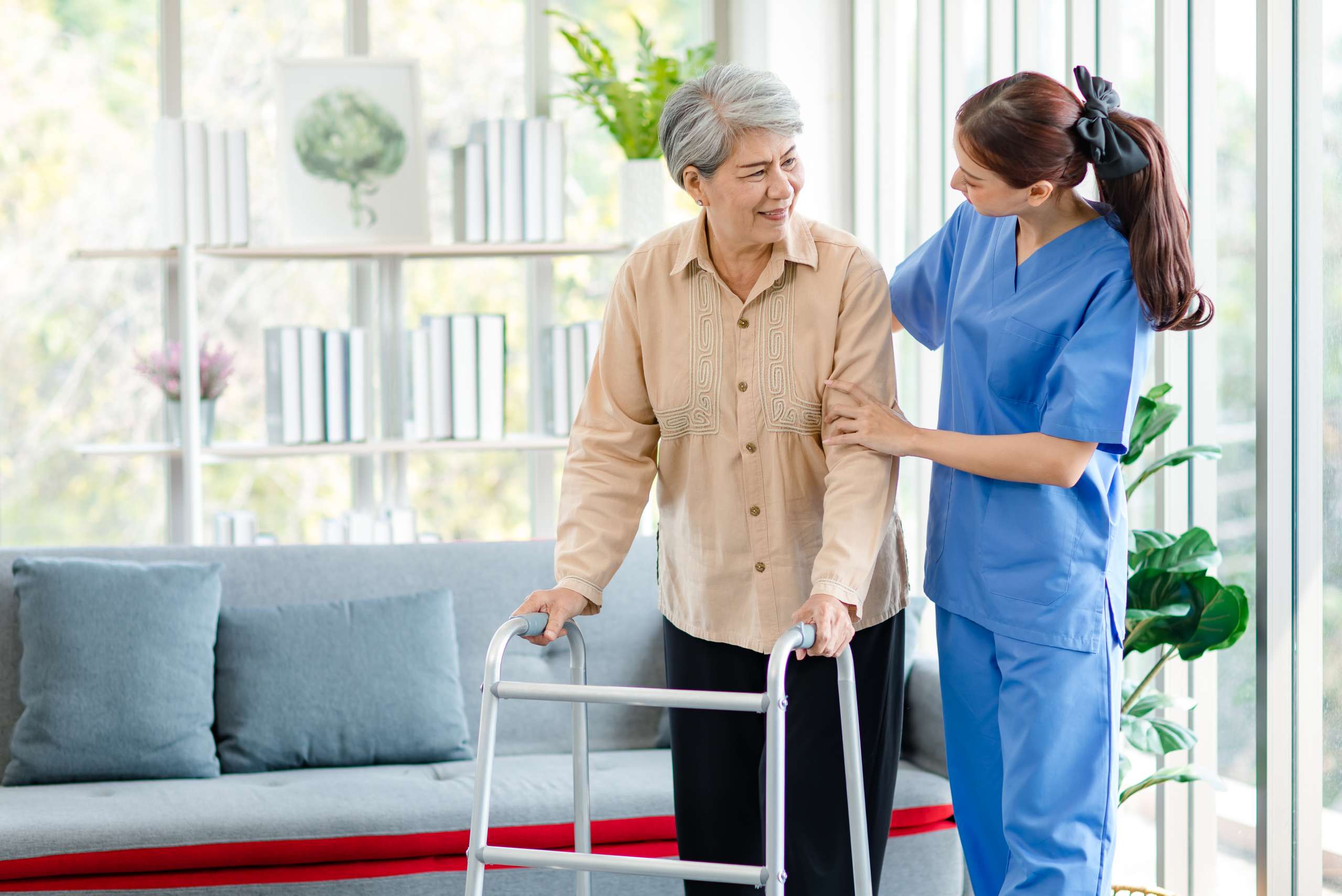 Young professional successful friendly female nurse in blue hospital uniform helping supporting physical therapy senior woman patient walking via four legs walker